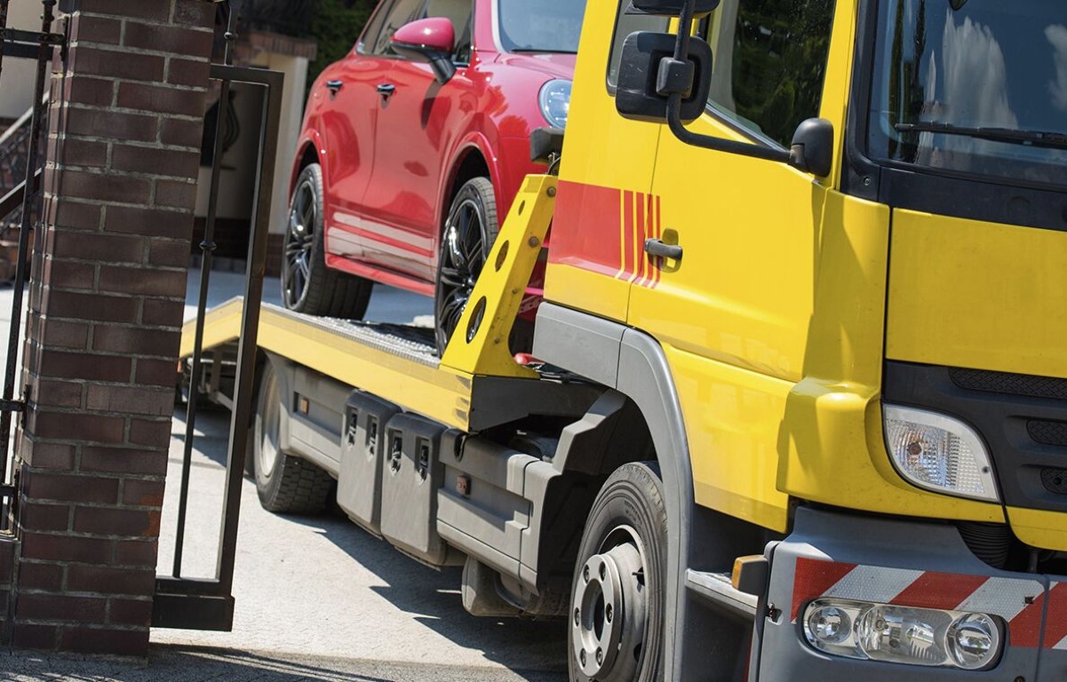 Yellow flatbed tow truck carrying red car through residential street in Lindenhurst, NY